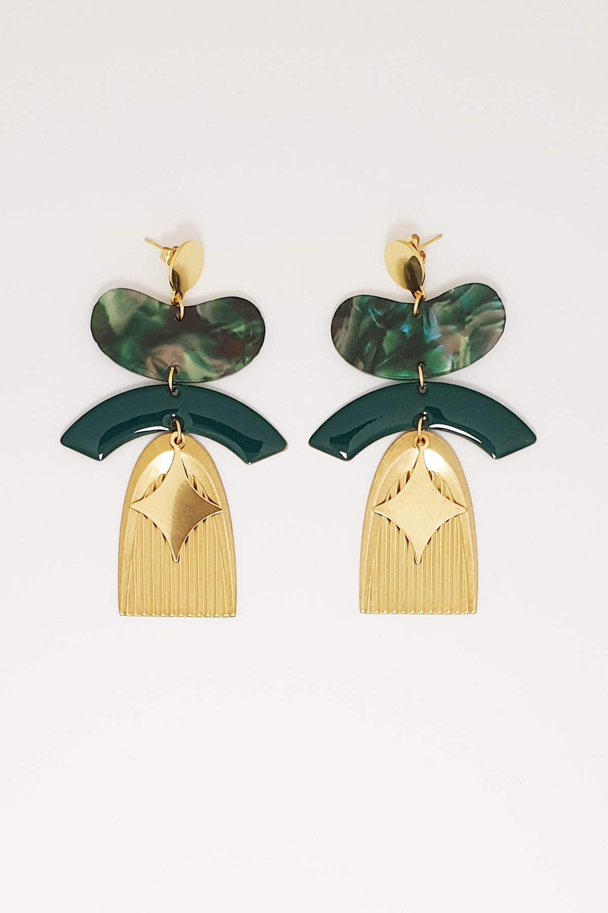 A pair of statement stud dangle earrings sit against a white background. They feature a green jellybean shaped acrylic piece, a green arch shaped enamel connector, a corrugated textured brass arch piece, and a diamond shaped brass piece.