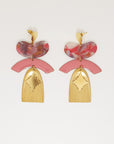 A pair of statement stud dangle earrings sit against a white background. They feature a pink jellybean shaped acrylic piece, a pink arch shaped enamel connector, a corrugated textured brass arch piece, and a diamond shaped brass piece. 