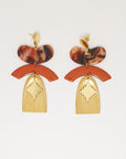 A pair of statement stud dangle earrings sit against a white background. They feature a tan jellybean shaped acrylic piece, a tan arch shaped enamel connector, a corrugated textured brass arch piece, and a diamond shaped brass piece.