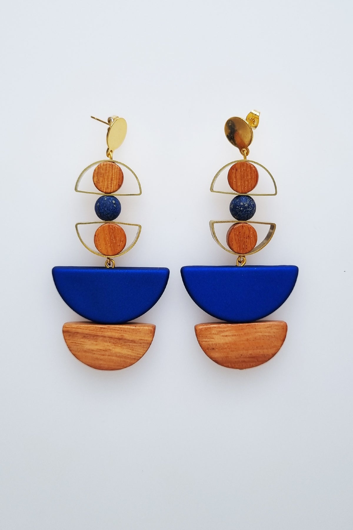 A pair of statement stud dangle earrings sit against a white background. They feature a small brass arch with a round wooden bead inside the arch, followed by a blue bead, another small brass arch upturned with a wooden circle bead inside, followed by a blue D shape bead, and wooden a D shape bead.