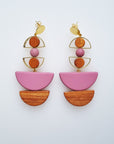 A pair of statement stud dangle earrings sit against a white background. They feature a small brass arch with a round wooden bead inside the arch, followed by a pink bead, another small brass arch upturned with a wooden circle bead inside, followed by a pink D shape bead, and wooden a D shape bead.