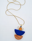 A necklace with a gold chain sits against a white background. It features a small wooden bead, followed by a brass D shape with a wooden circle bead enclosed, followed by a blue D shaped bead, and a wooden D shape bead.