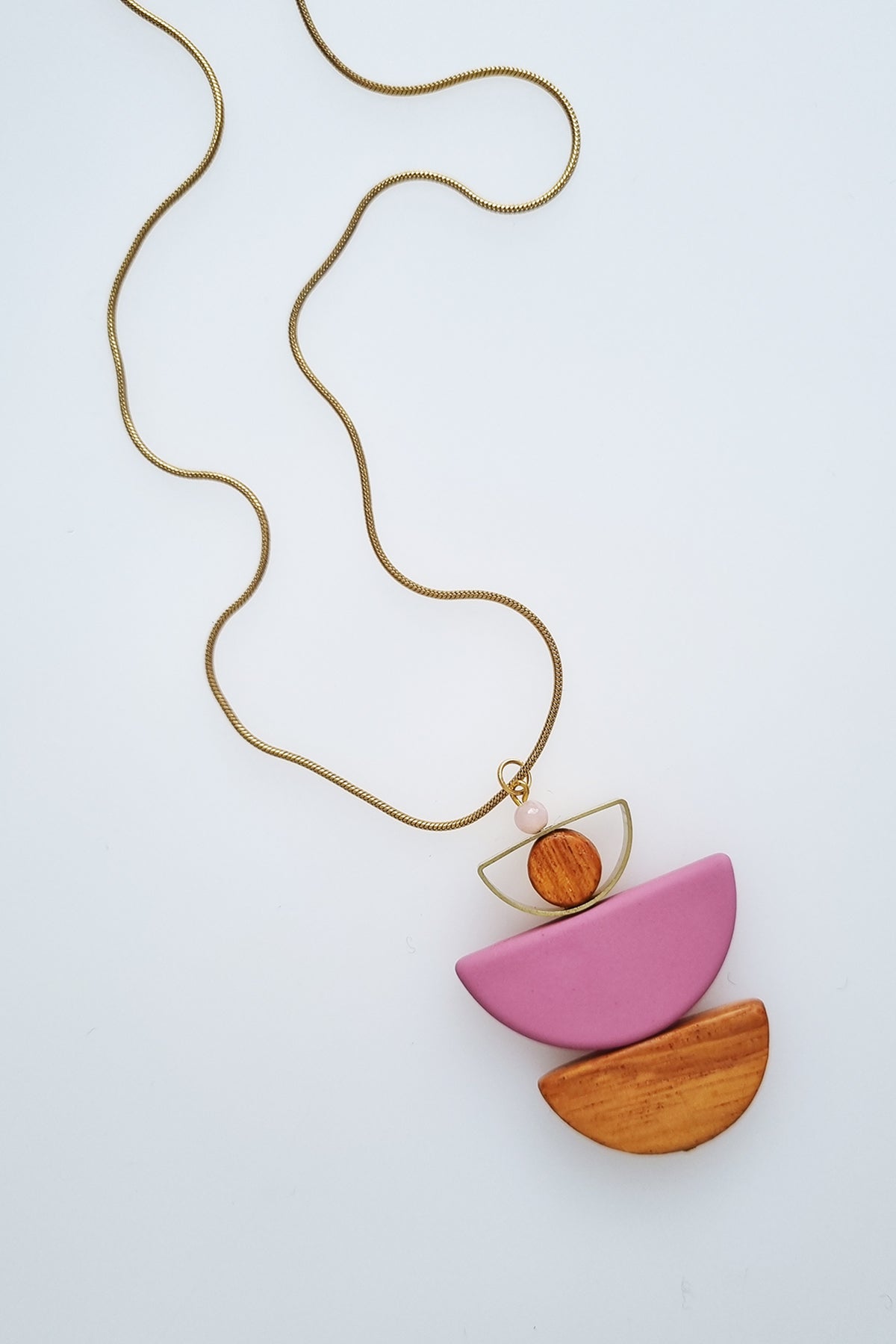 A necklace with a gold chain sits against a white background. It features a small wooden bead, followed by a brass D shape with a wooden circle bead enclosed, followed by a pink D shaped bead, and a wooden D shape bead.