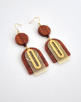 A pair of dangle earrings with a hook sit against a white background. They feature a circular wooden bead with a tiny terracotta bead. Attached below this is a terracotta arch shaped acrylic piece, an arch shaped brass piece, and an elongated oval brass piece.