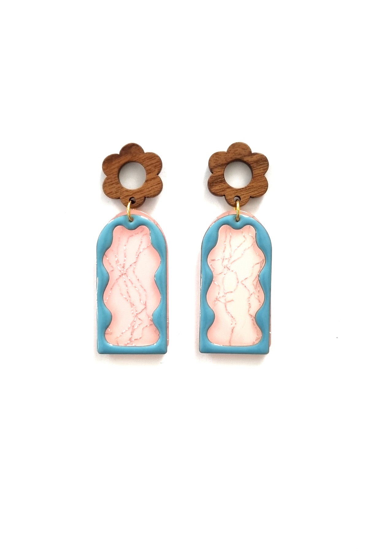 A pair of stud dangle earrings lay against a white background. They feature a wooden flower stud top followed by a pink acrylic arch with a silver thread detail. On top sits an enamel wavy arch frame in blue.