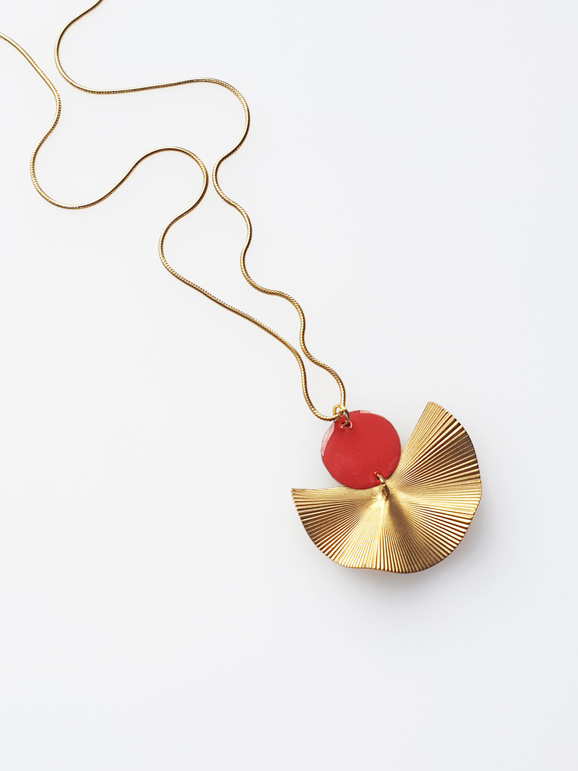 A necklace sits against a white background. It features a gold chain, a coral enamel connector dot and a textured brass fan piece.