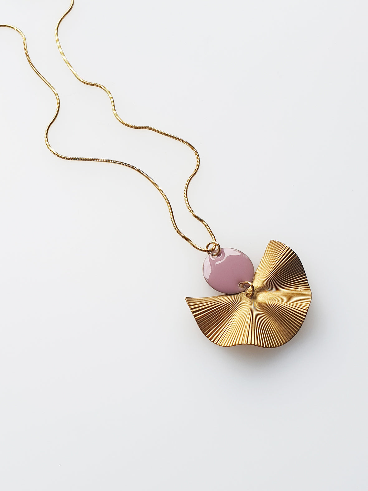 A necklace sits against a white background. It features a gold chain, a mauve enamel connector dot and a textured brass fan piece.