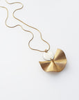 A necklace sits against a white background. It features a gold chain, a white enamel connector dot and a textured brass fan piece.