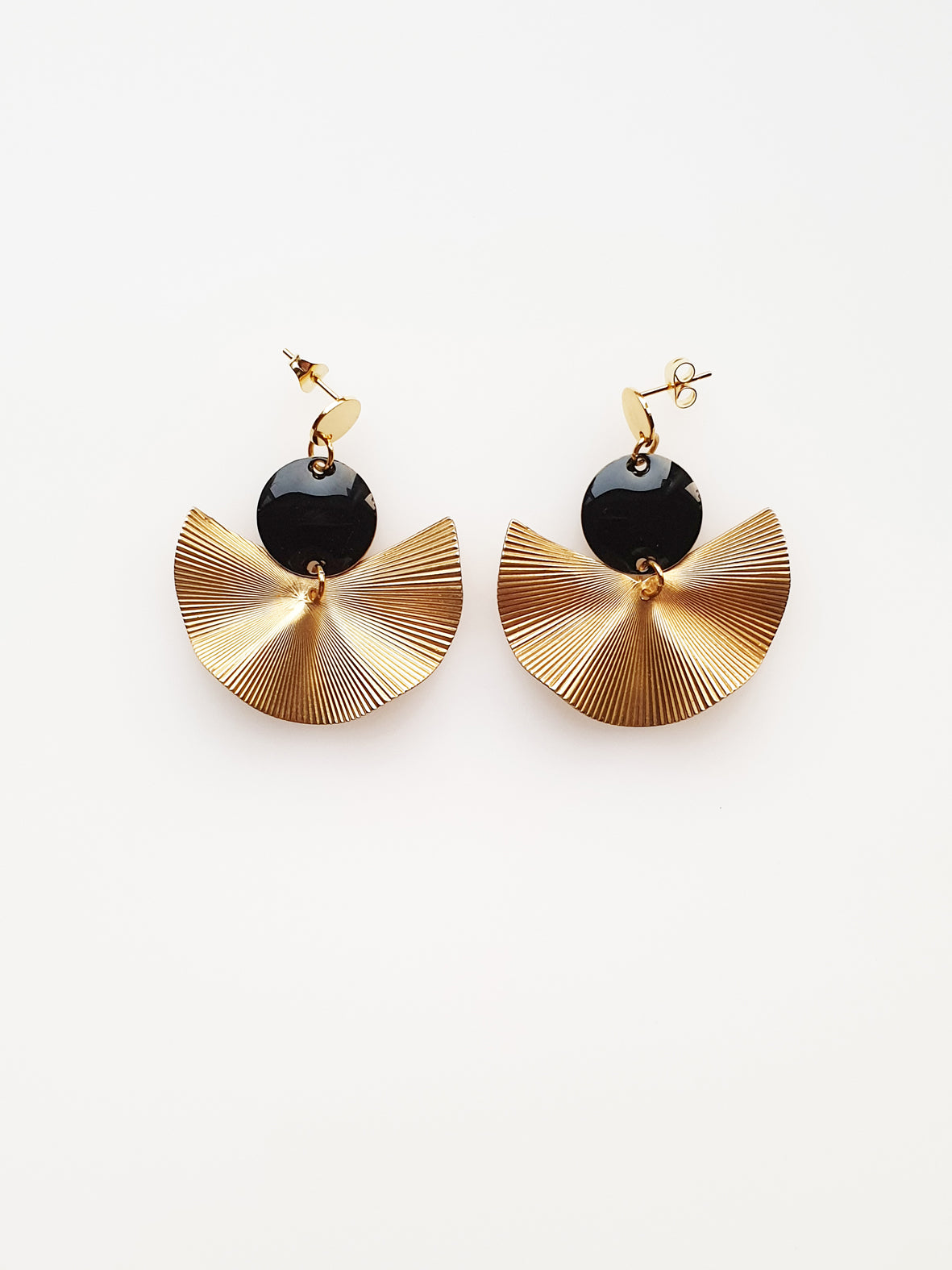 A pair of stud dangle earrings sit against a white background. They feature a black enamel connector dot and a textured brass fan piece.