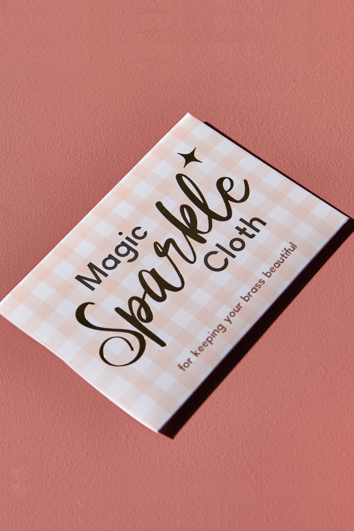 An pink checkered envelope with the words 'Magic Sparkle Cloth for keeping your brass beautiful' written in gold foil, sits against a pink background.