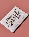 An pink checkered envelope with the words 'Magic Sparkle Cloth for keeping your brass beautiful' written in gold foil, sits against a pink background.