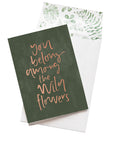 A dark green card with the message 'you belong among the wild flowers' is sitting on a white background. The card uses rose gold script font. There is a white envelope behind the card which has a green watercolour plant design inside.