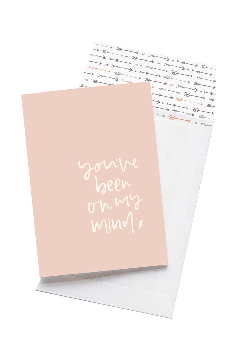 A pale pink card with the message &#39;you&#39;ve been on my mind x&#39; is sitting on a white background. The card uses white script font. There is a white envelope behind the card which has an arrow pattern design inside.